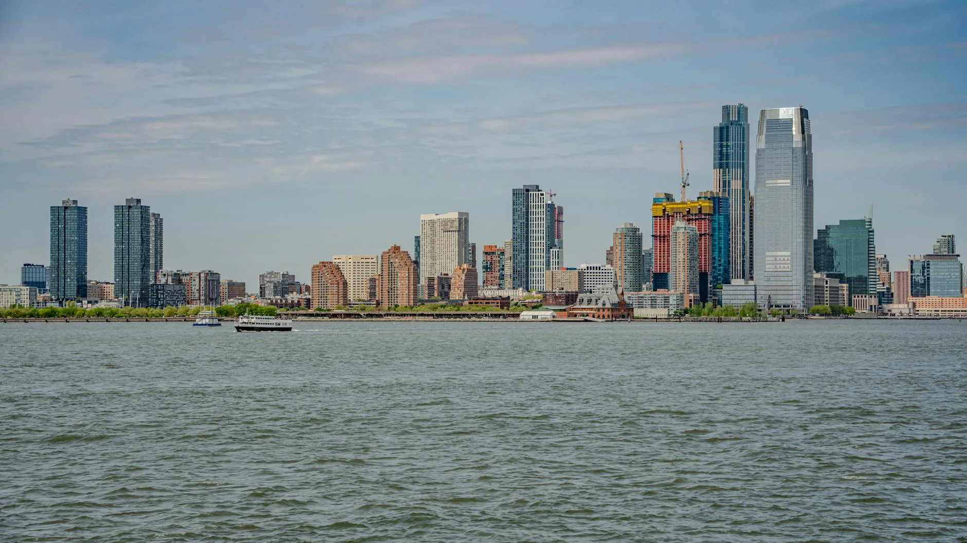 The Jersey City skyline viewed across the Hudson River, with high-rise buildings and active construction along the waterfront